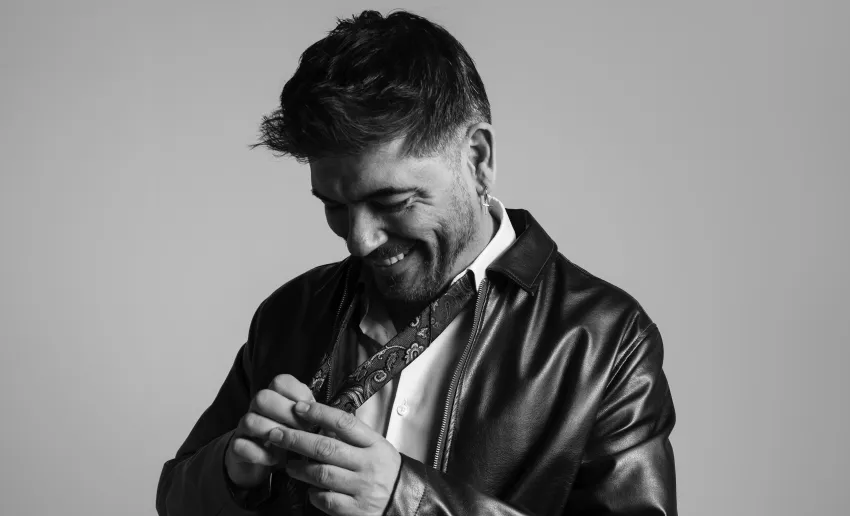 foto en blanco y negro, hombre mirandose hacia abajo sus propias manos que tocan los extremos del pañuelo que lleva al cuello, chaqueta de cuero, camisa blanca