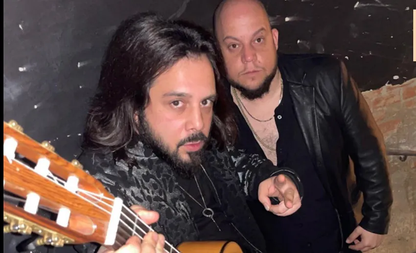 dos hombres con barba y perilla, mirando a cámara con gesto serio, uno de ellos sostiene  una guitarra y hace el gesto de la v con dos dedos de la otra mano
