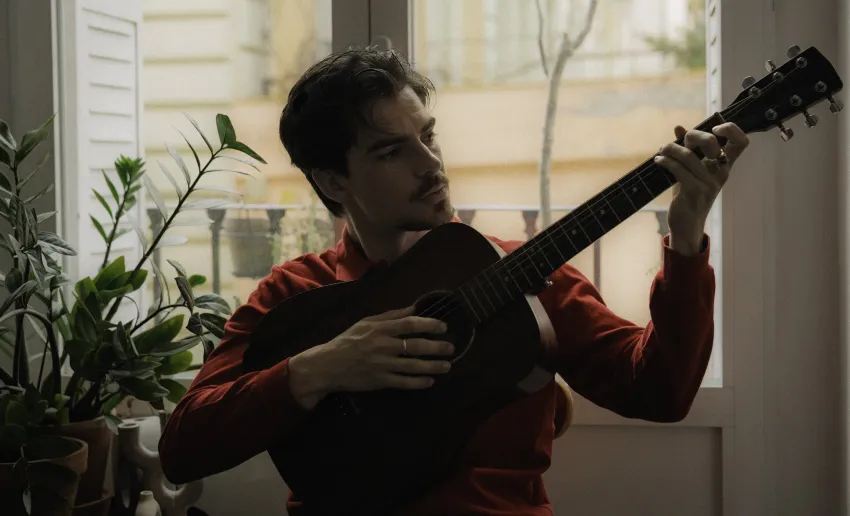 hombre tocando la guitarra, a su espalda una ventana y plantas