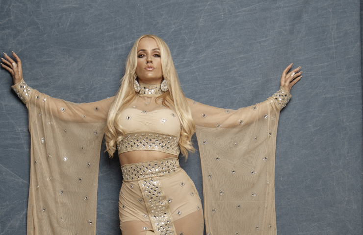 Mujer rubia vestida de dorado frente a fondo de tela gris