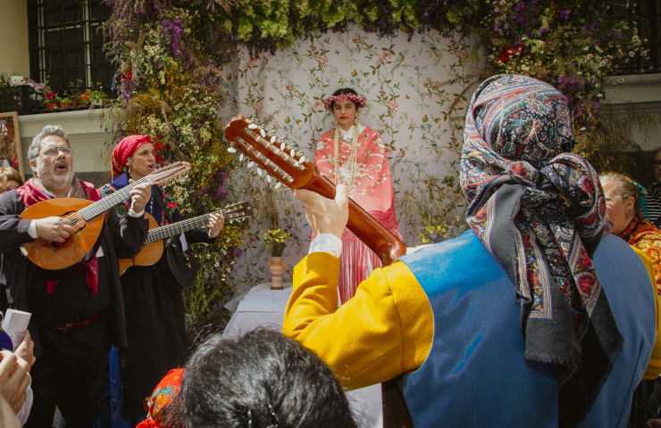 personas con instrumentos musicales con trajes regionales cantándole a una niña sentada con mantón sobre los hombros y corona de flores