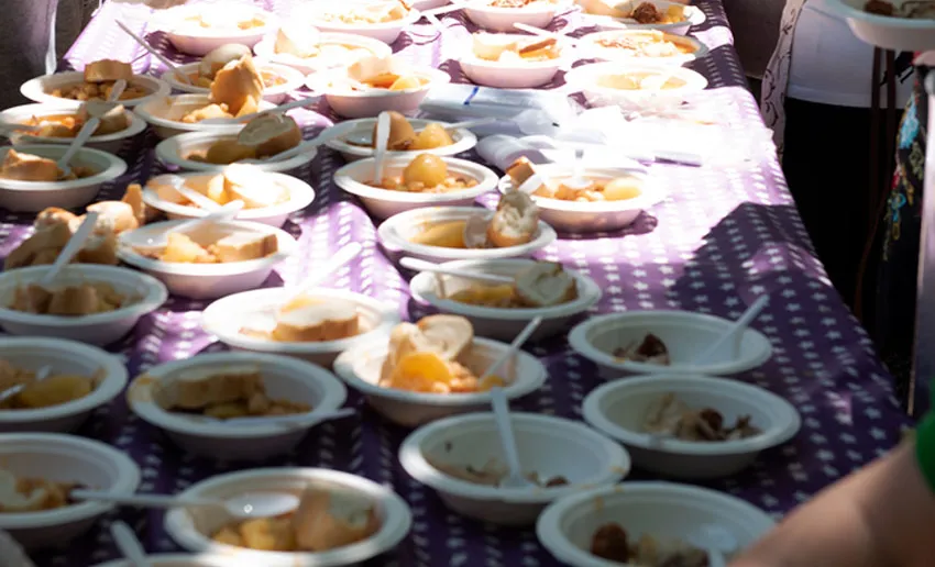 mesa con mantel de cuadros, llena de platos de plastico con cocido, un trozo de pan y un cubierto