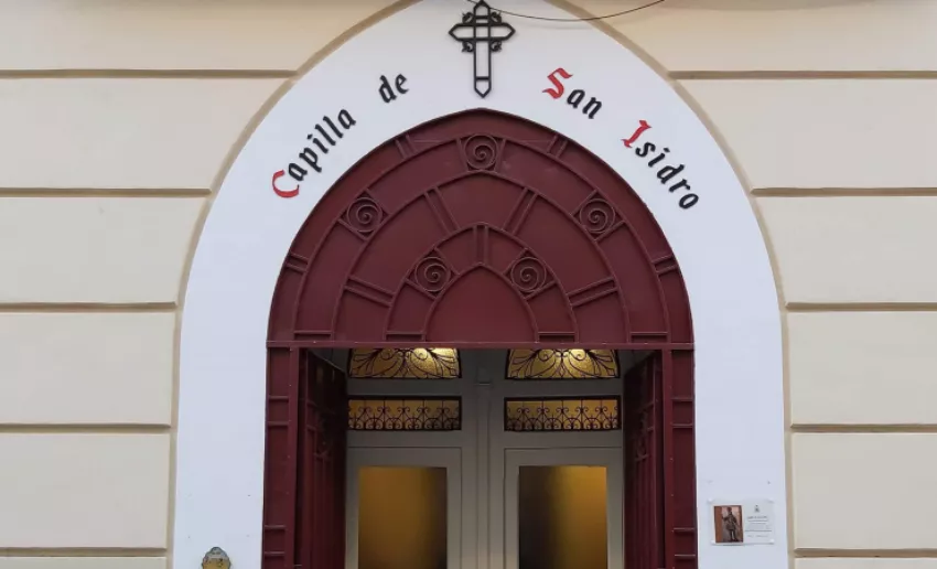 Entrada de la Capilla de San Isidro, con puerta doble de madera enmarcada por un arco decorativo en tonos rojizos y el nombre de la capilla rotulado en la parte superior de la fachada.