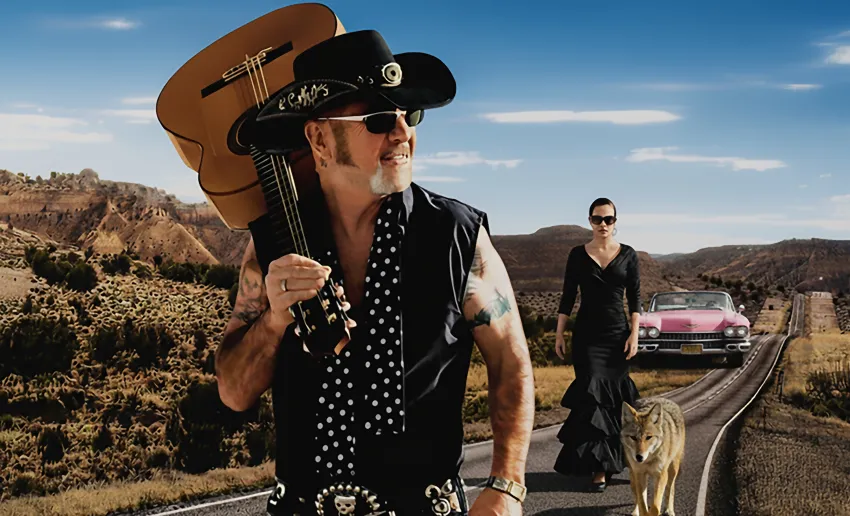 hombre con grandes patillas, gorro negro de cowboy, chaleco negro, gafas de sol, guitarra apoyada al hombro, brazos tatuados, cinturón con una gran hebilla metálica y pañuelo de lunares. mujesr en segundo plano con traje de flamenca en negro.