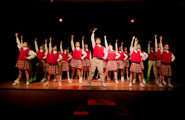 coro de niños y niñas sobre escenario, todos vestidos igual chaleco rojo, camisa blanca, pantalón beige o falda de cuadros beige y rojos
