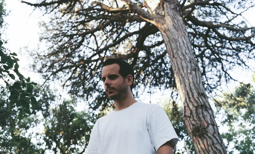 hombre en medio de la naturaleza, rodeado de árboles, mirada hacia abajo, camiseta blanca de manga corta
