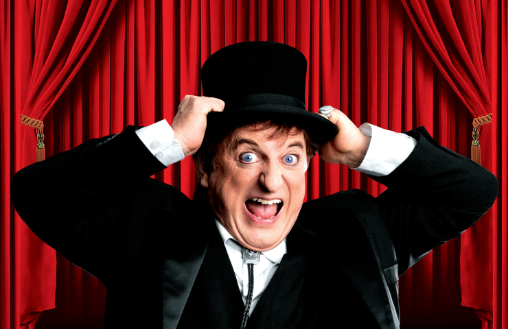 Hombre vestido de negro, frente a unas cortinas rojas, sujetándose su chistera