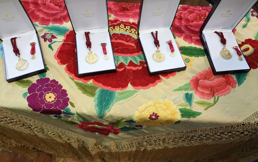 4 medallas doradas con cordón rojo, dentro de unas cajas cuyo interior es blanco, sobre una mesa cubierta por un mantón de manila color beige con grandes flores bordadas de diferentes colores